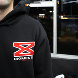 Person wearing a black hoodie with a red and white logo on the back, standing indoors.
